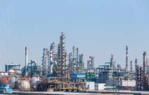 A wide view of an industrial complex featuring various refinery structures, including tall towers, storage tanks, and pipelines, against a clear blue sky, representing the oil and petrochemical industries.