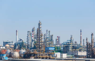 A wide view of an industrial complex featuring various refinery structures, including tall towers, storage tanks, and pipelines, against a clear blue sky, representing the oil and petrochemical industries.