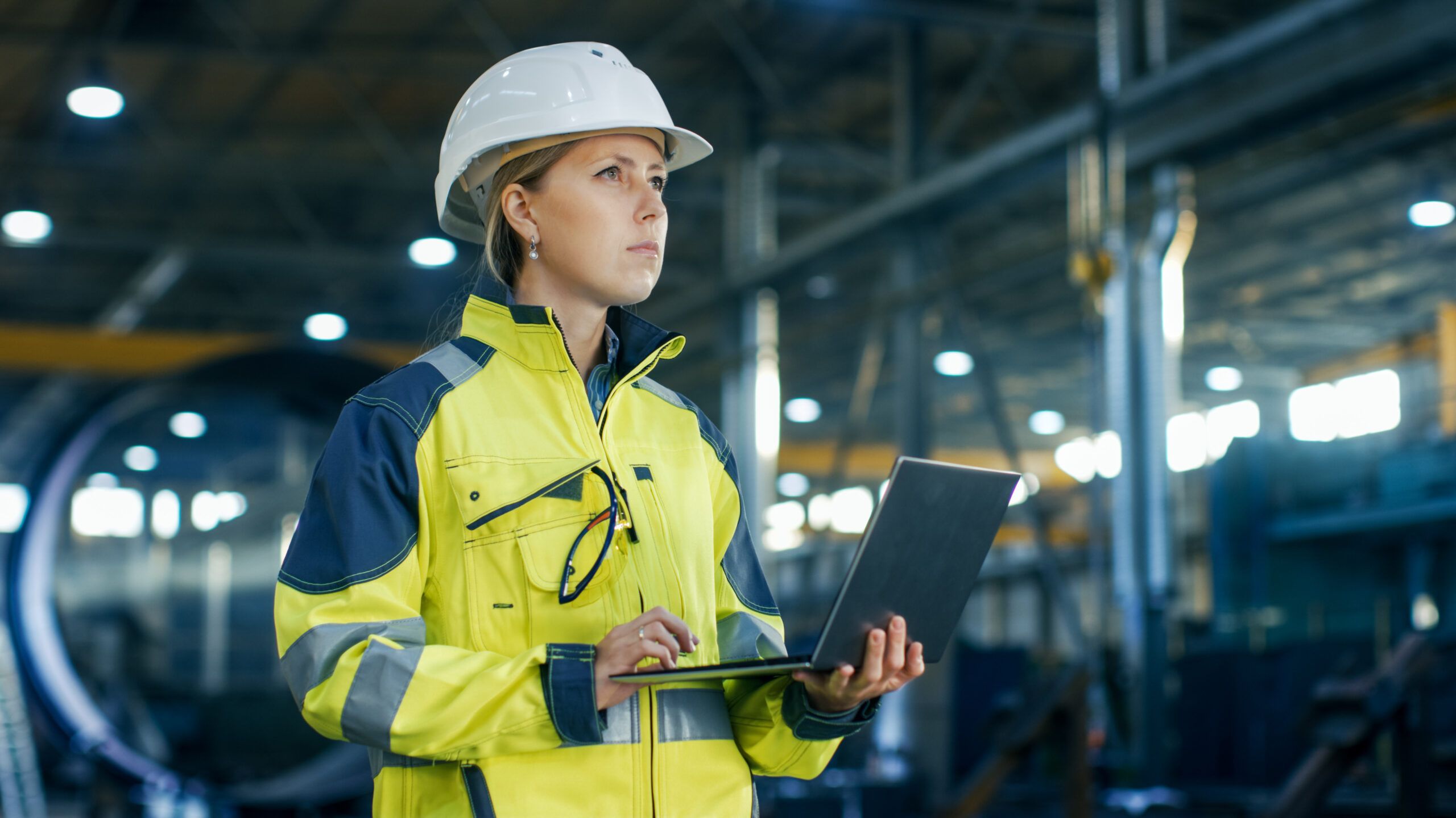A female engineer wearing a safety helmet and a high-visibility jacket stands in an industrial setting, holding a laptop and looking thoughtfully. The background shows a large, well-lit workspace, indicating a focus on engineering and digital transformation.