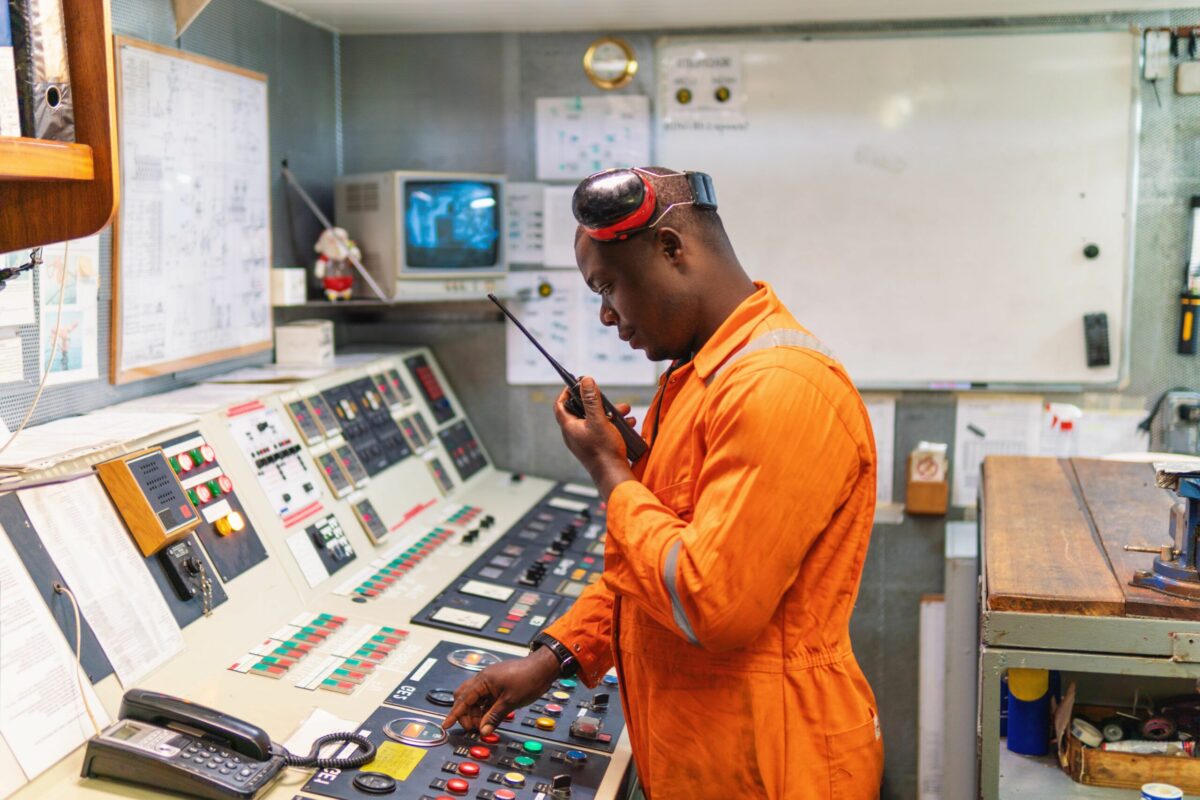 Engineer in orange coveralls using radio communication while operating an industrial control panel inside a control room.