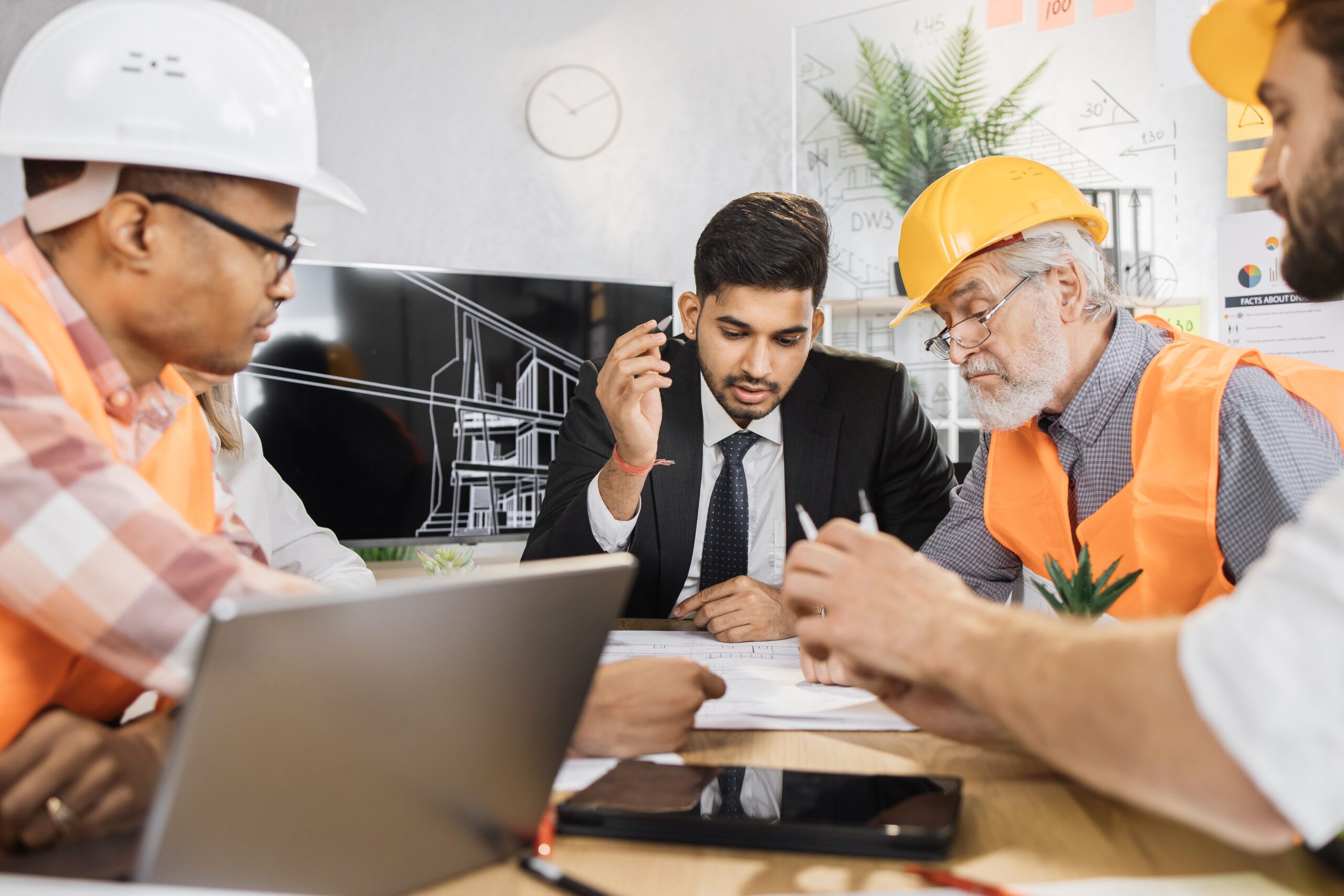 Electrical engineers and a project manager review plans and discuss interview questions during a collaborative technical meeting.