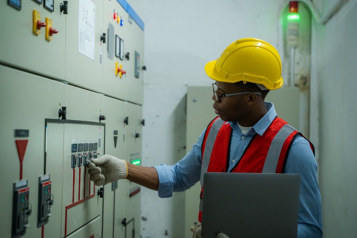 Engineer in safety gear inspecting a high-voltage electrical control panel while holding a laptop.