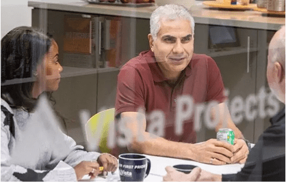 A diverse team engaged in a meeting at Vista Projects. One participant holds a drink, while another takes notes. The focus is on collaboration and discussion regarding engineering solutions.