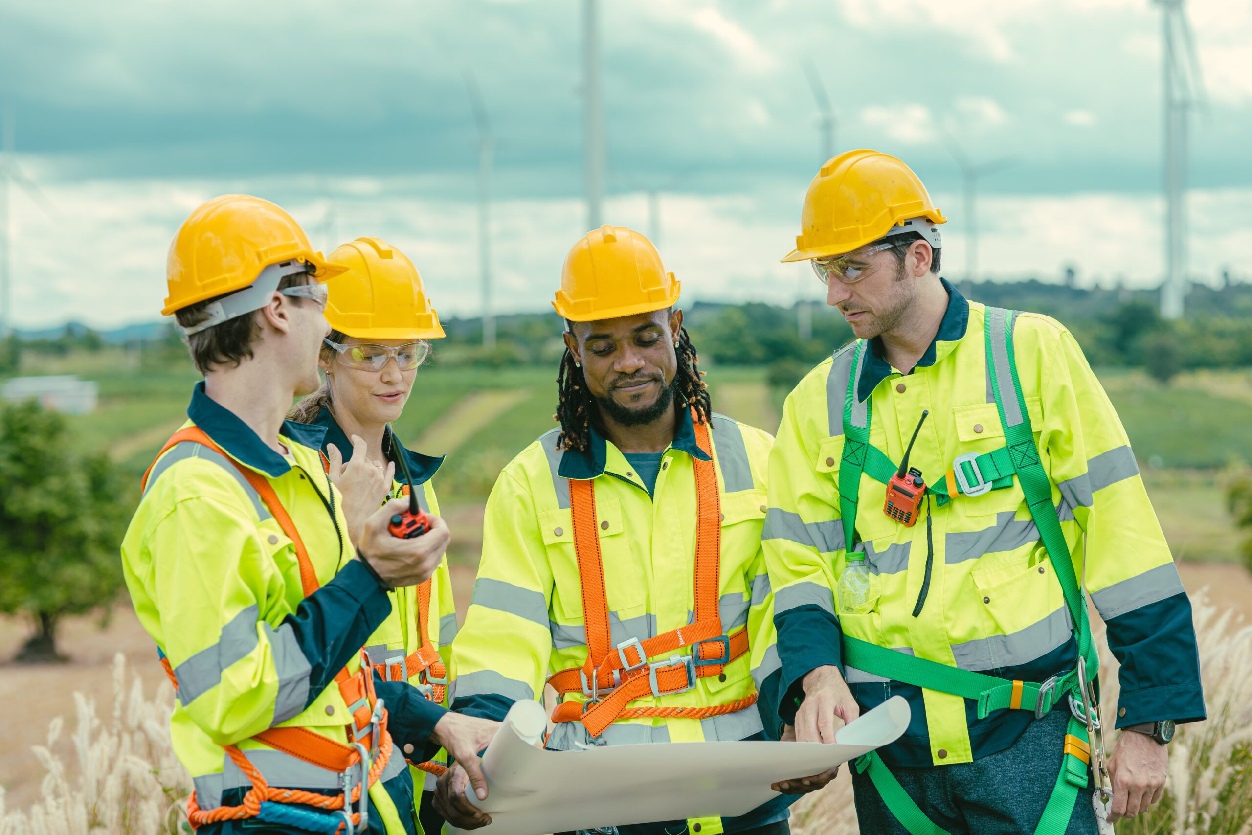 Four engineering professionals in safety gear discuss plans in a field with wind turbines in the background. They are reviewing technical drawings and collaborating on a project, highlighting teamwork in engineering and technical fields.
