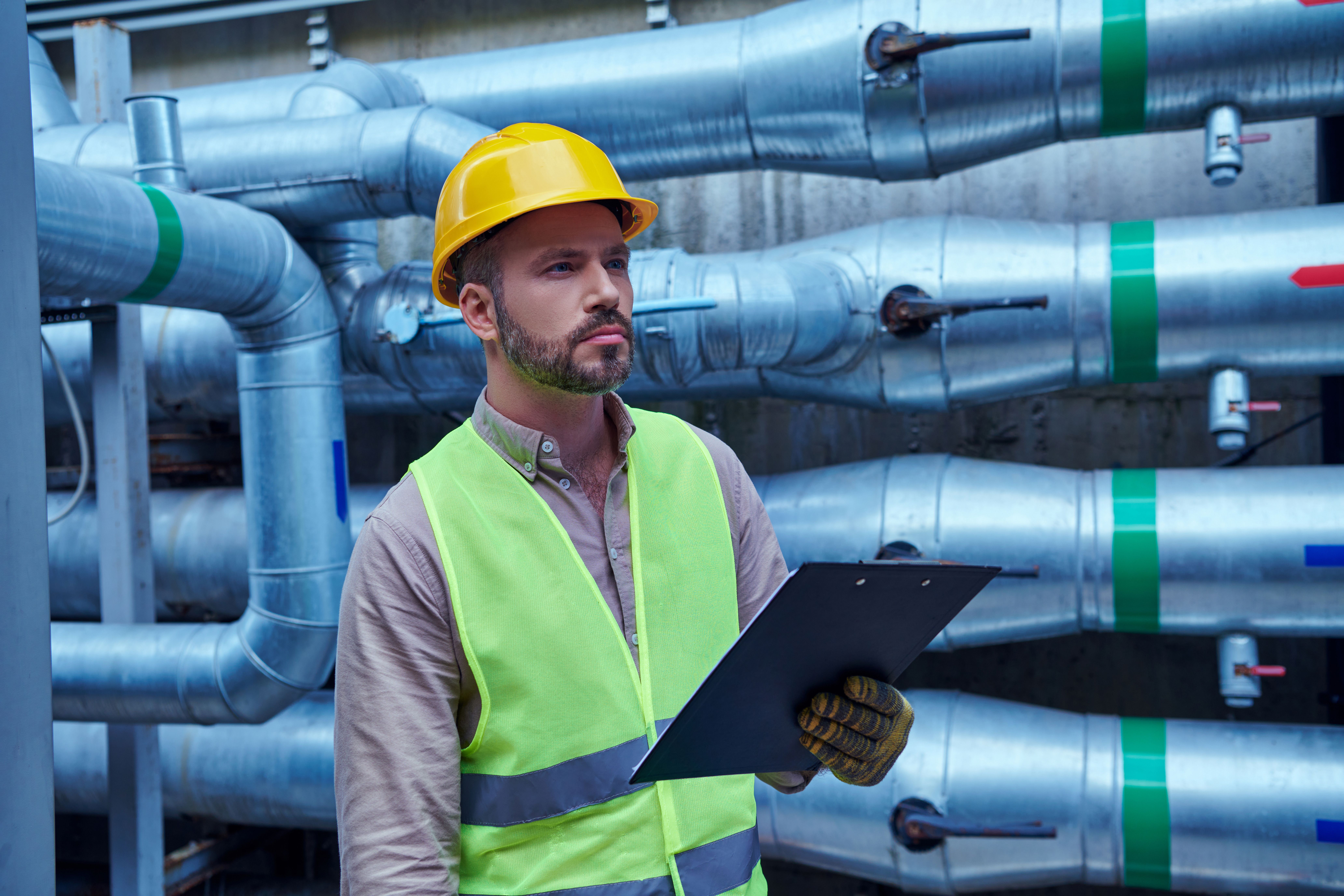 Industrial piping inspector in safety gear evaluates complex pipe system with color-coded bands in a mechanical facility.