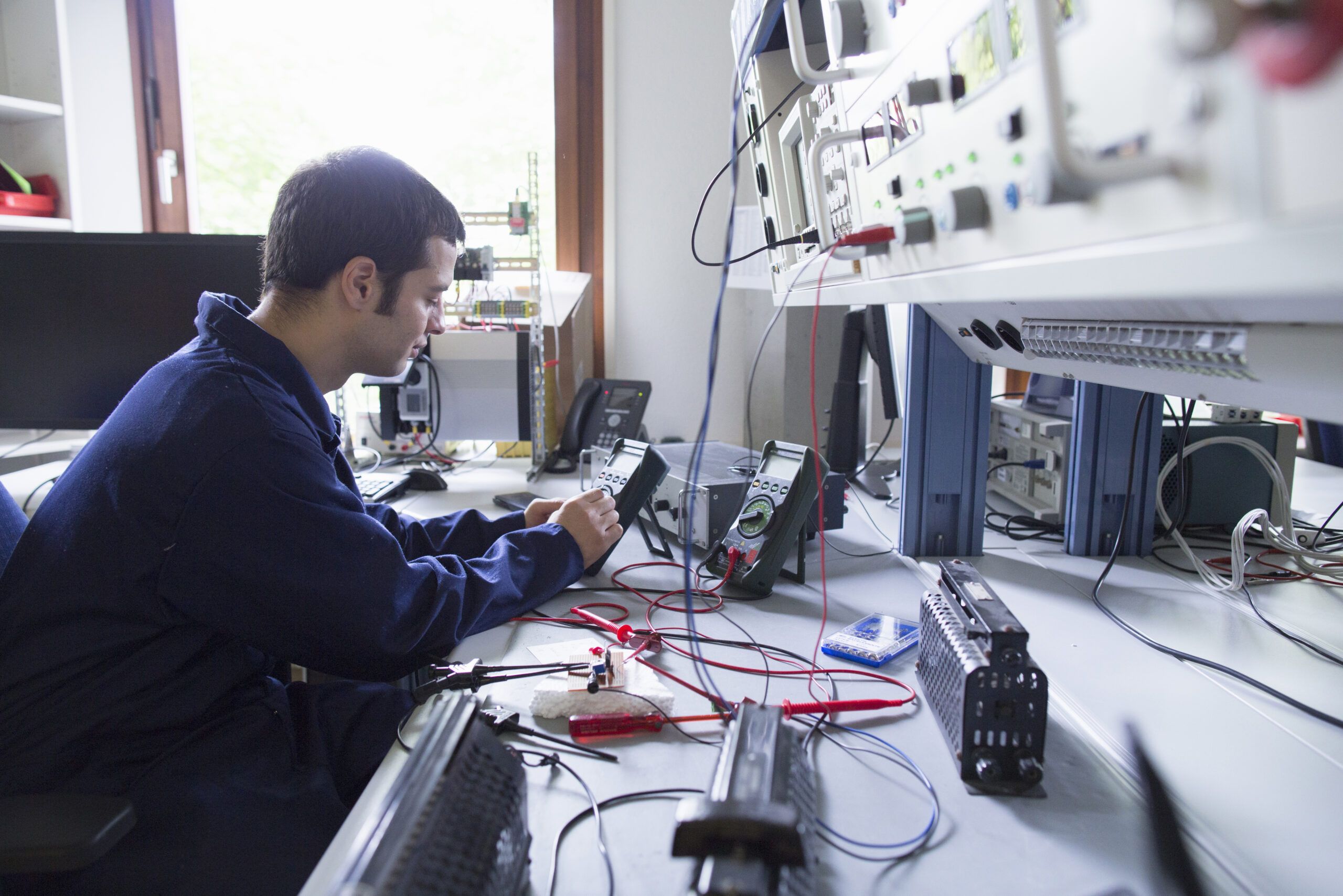 A technician performing intricate, close-up electronic testing on a control system component or instrument at a workbench with specialized test equipment like multimeters.